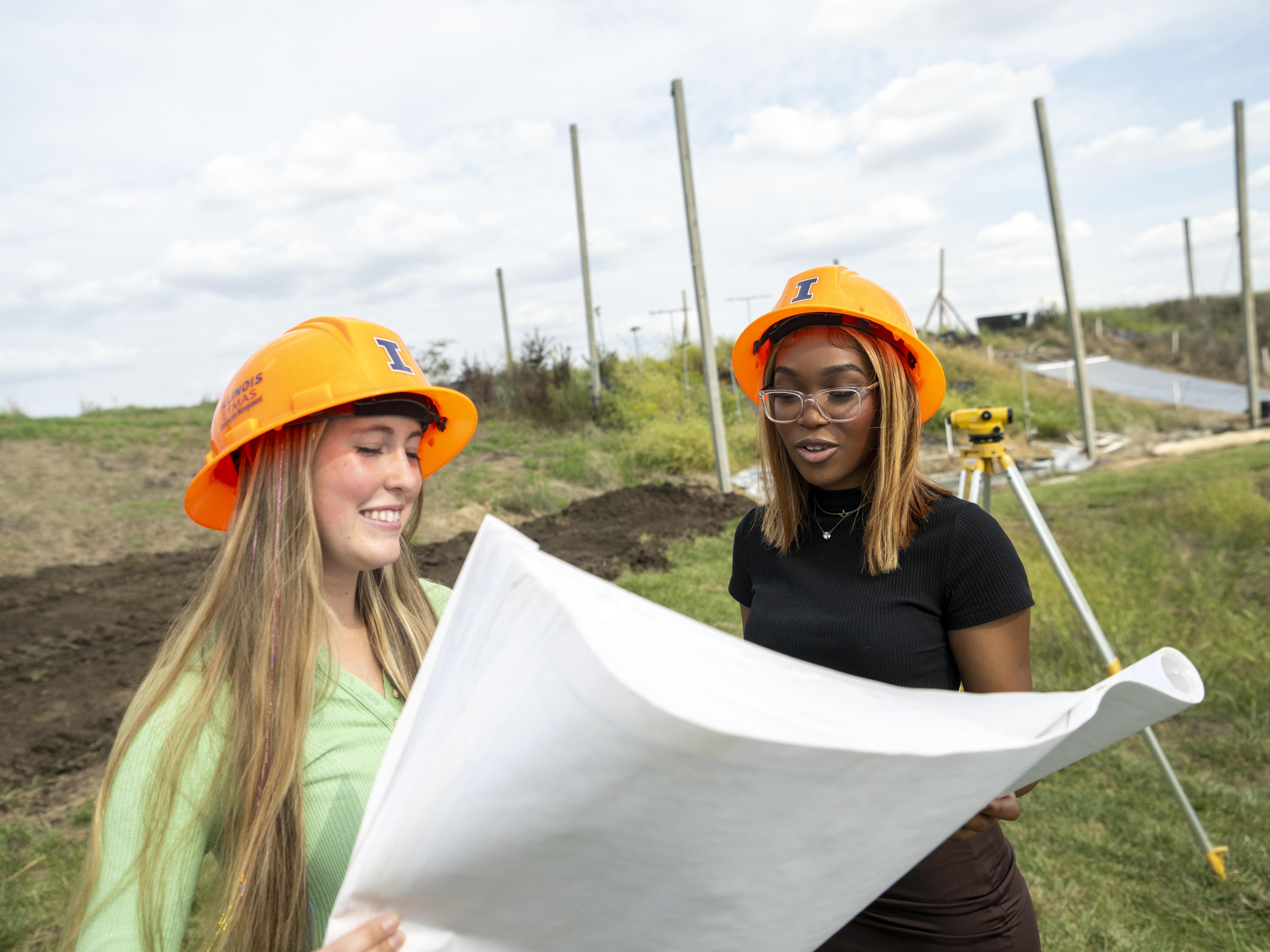 Two women in hard hats looking at a blueprint in an agricultural field.